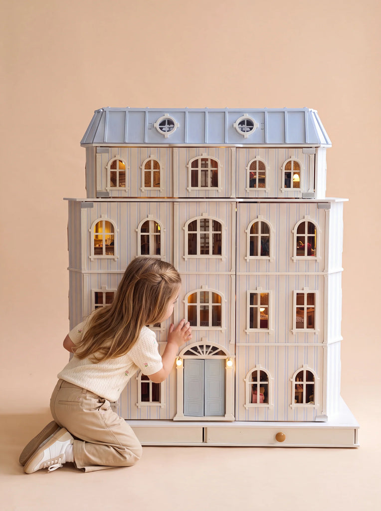 A young child kneels and peers inside the Fully Furnished Dollhouse With Lights, House of Monaco — The Grand Mansion, its illuminated windows glowing against a plain beige background.