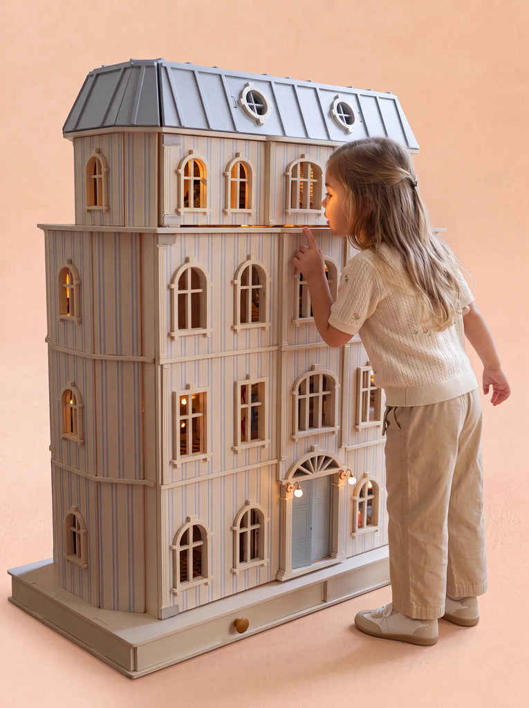 A young girl in a white shirt and beige pants gazes into the upper window of the House of Monaco — The Grand Mansion: a large, fully furnished dollhouse with glowing lights inside, set against a plain peach background.