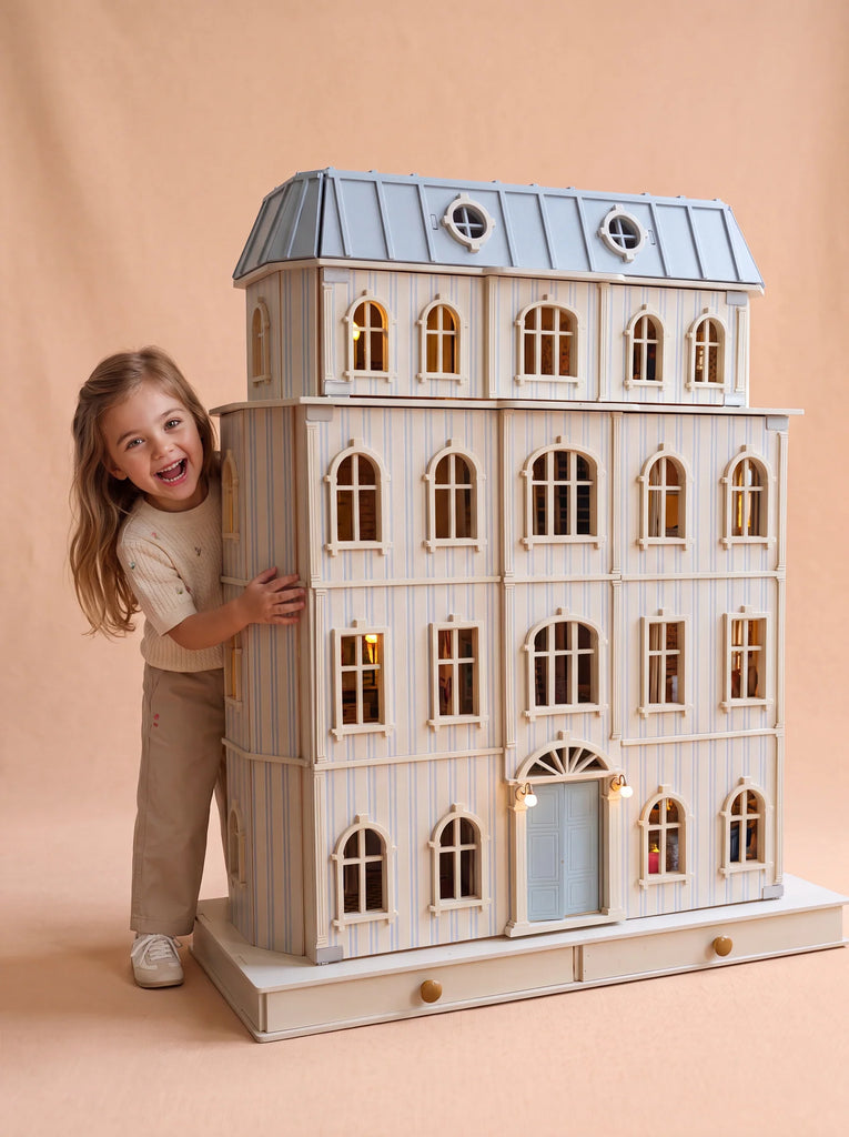 A smiling child in beige clothes stands beside the Fully Furnished Dollhouse With Lights, House of Monaco — The Grand Mansion, a large and intricately detailed luxury dollhouse featuring blue and white stripes and arched windows, set against a plain tan background.