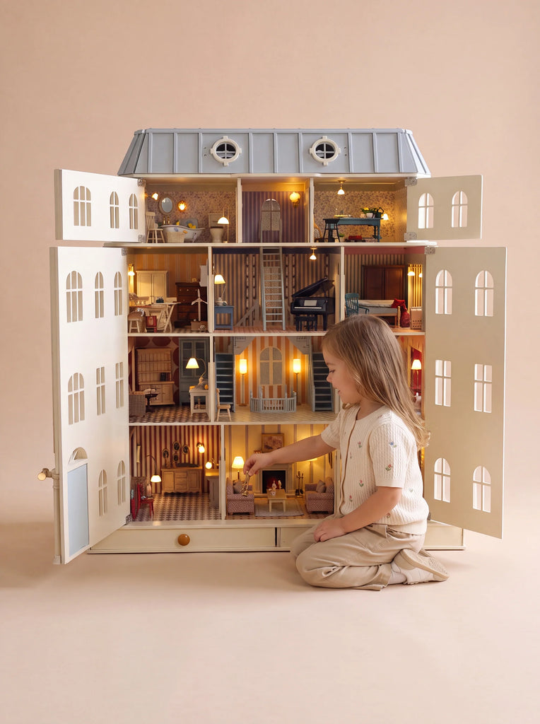 A young child sits cross-legged and plays with the Fully Furnished Dollhouse With Lights, House of Monaco — The Grand Mansion, a spacious, beautifully illuminated Haussmann-inspired dollhouse featuring multiple rooms and intricate details.