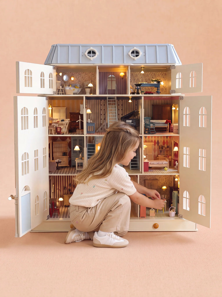 A young child kneels and plays with the House of Monaco — The Grand Mansion, a fully furnished dollhouse with lights featuring illuminated rooms and miniature furniture inspired by Haussmann-style elegance, all set against a plain beige background.