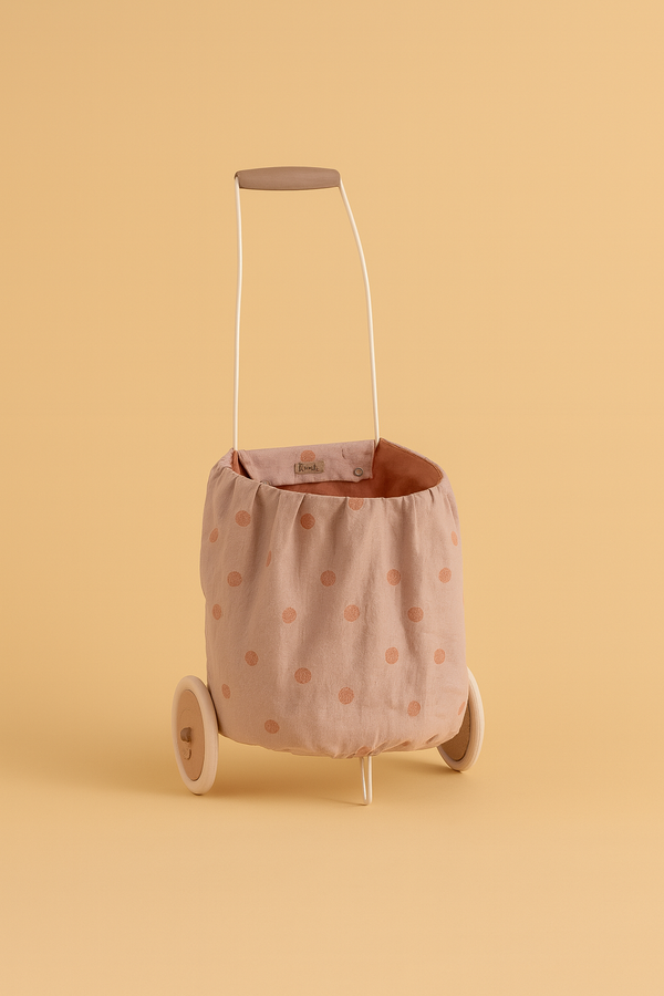 The Maileg Trolley, Multi dots - Rose is a small, washable cart with a metal handle and beige linen body covered in pink polka dots, displayed against a solid peach backdrop.