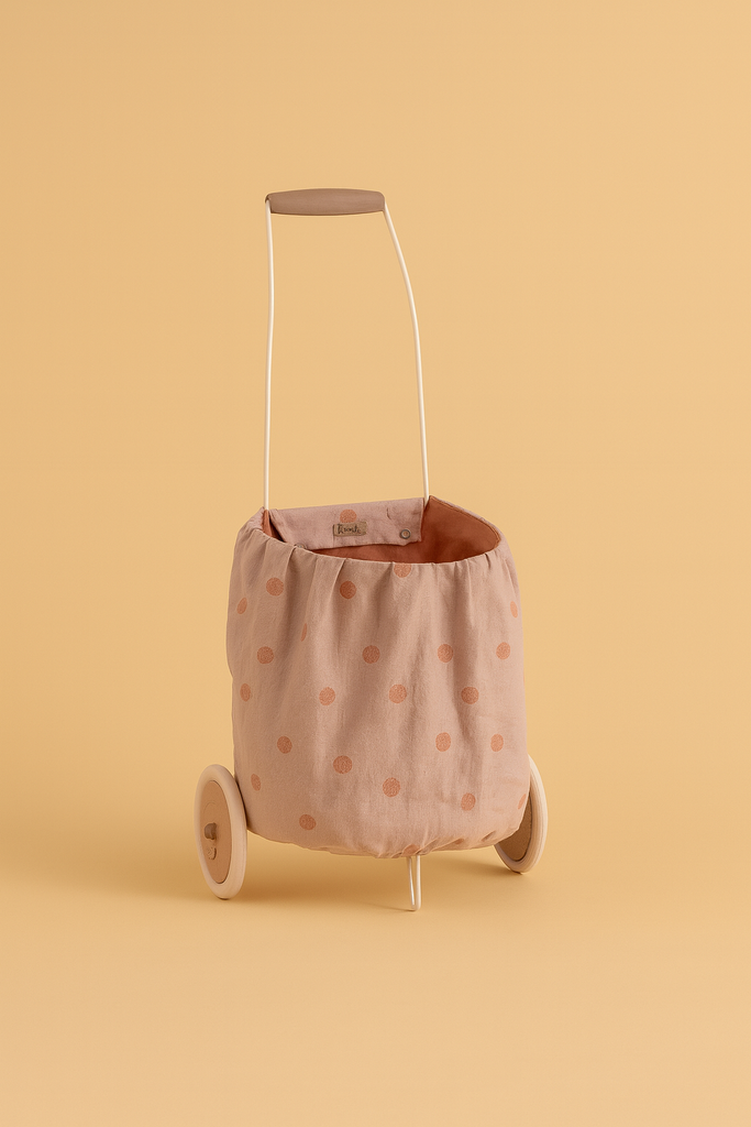 The Maileg Trolley, Multi dots - Rose is a small, washable cart with a metal handle and beige linen body covered in pink polka dots, displayed against a solid peach backdrop.