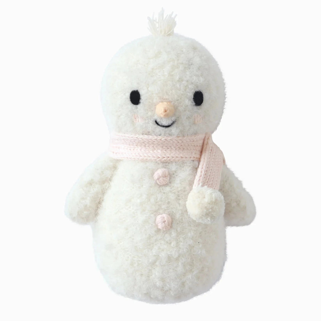 The Cuddle + Kind Baby Snowman - Blush is a fair trade plush toy featuring a soft, fuzzy texture, black eyes, an orange nose, and a pale pink scarf—ideal for stuffed animal lovers and winter decor.