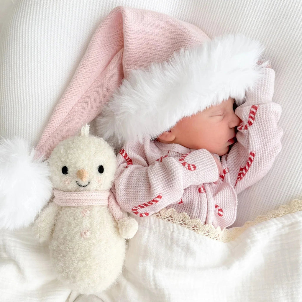 A newborn sleeps peacefully in a pink Santa hat and candy cane outfit, cuddled up with the Cuddle + Kind Baby Snowman - Blush plush under a cozy white blanket, surrounded by fair trade stuffed animals.