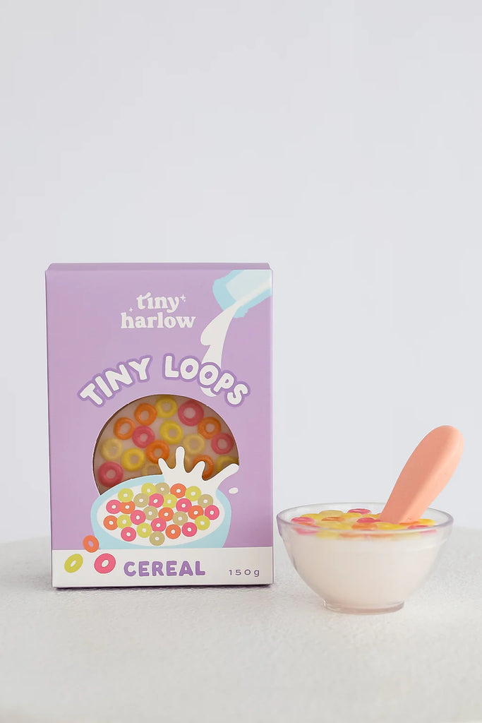 A purple box labeled "Tiny Harlow Tiny Loops Cereal" stands beside a Tiny Tummies - Tiny Loops Cereal Bowl filled with colorful loops and a pink spoon, perfect for doll breakfast play, set against a simple light background.