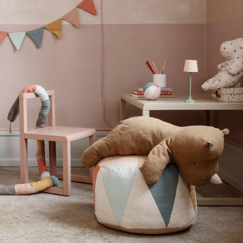 A cozy children's room with pastel decor, featuring a stuffed bear on the Maileg Drum Pouf, a pastel chair with a soft toy, a small table with a lamp and toys, and a colorful pennant banner on the wall.