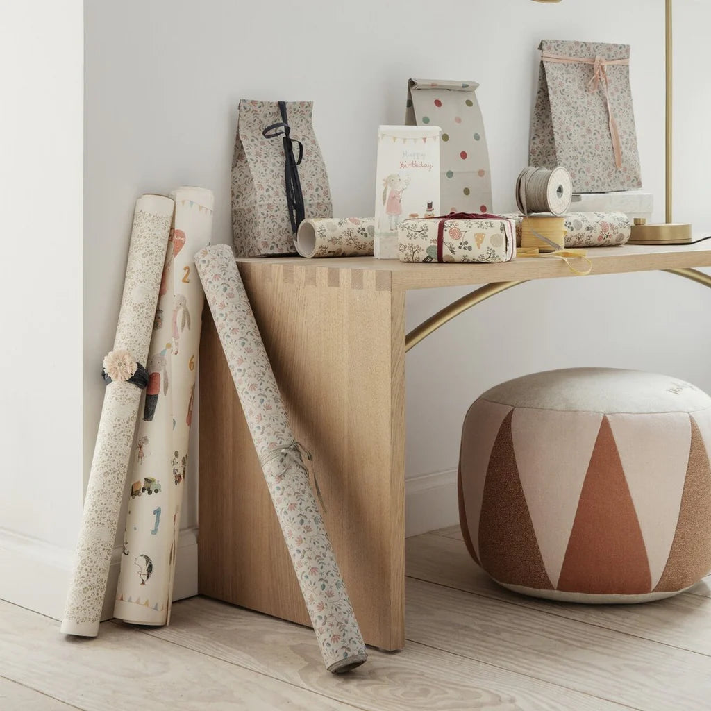 A modern room displays decorative paper, gift bags, a birthday card, and ribbon spools on a wooden desk beside a Maileg Drum Pouf with a geometric pattern on a light wood floor.