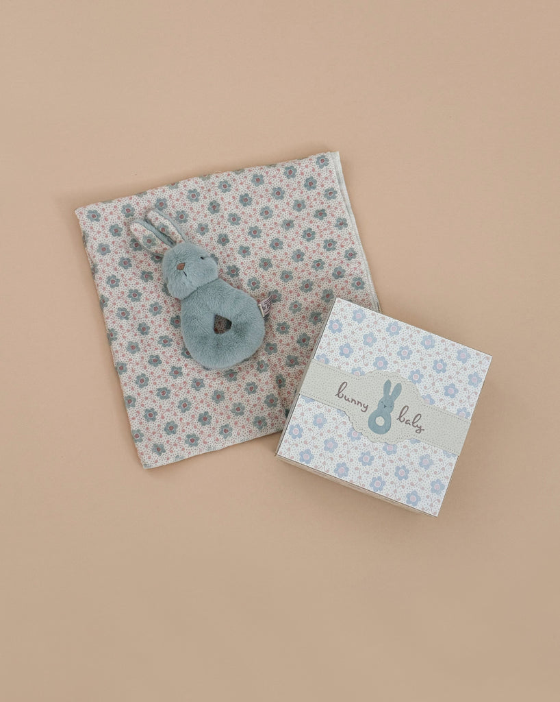 A soft gray bunny rattle sits on patterned cloth next to a matching "bunny baby" box with a bunny design and the Grimm's Small Wooden Rainbow, all part of the Newborn Gift Set - Blue, set against a beige background.