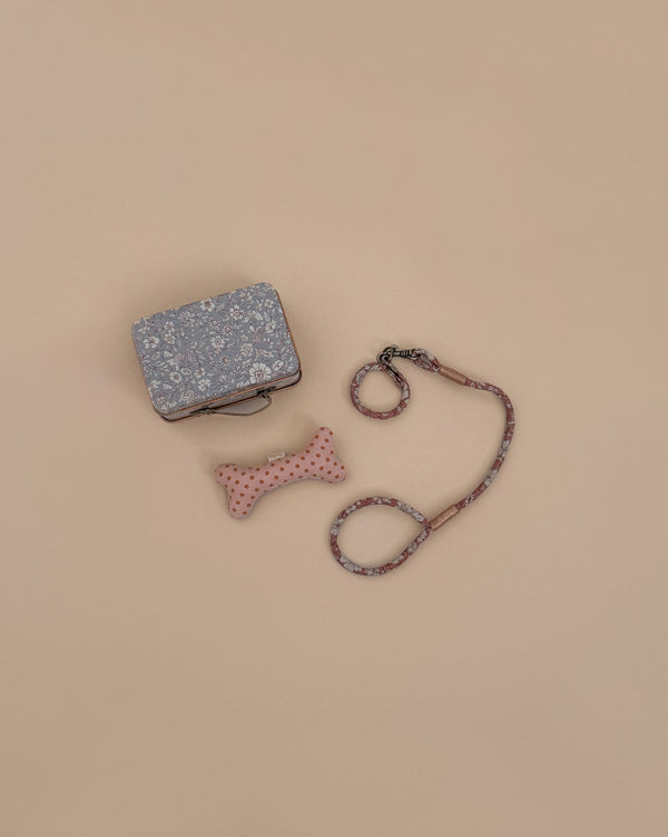 The Maileg Puppy Supply Accessories - Rose, featuring a floral-patterned bag, pink polka-dotted bone toy, and matching leash, are displayed on a beige background.