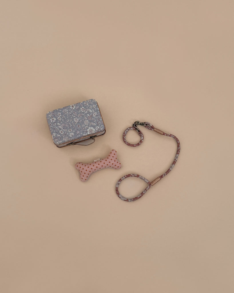The Maileg Puppy Supply Accessories - Rose, featuring a floral-patterned bag, pink polka-dotted bone toy, and matching leash, are displayed on a beige background.