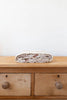 The Organic Cotton Baby Blanket - Pony, in beige and brown with a crochet trim, is neatly folded atop a two-drawer wooden dresser against a plain white wall.