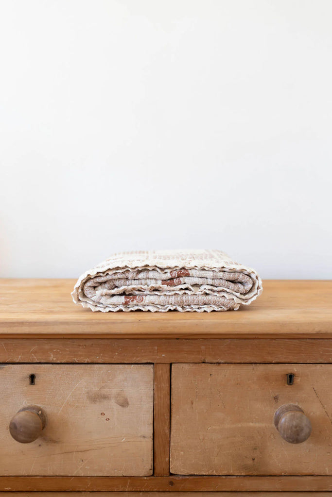 The Organic Cotton Baby Blanket - Pony, in beige and brown with a crochet trim, is neatly folded atop a two-drawer wooden dresser against a plain white wall.