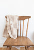 A brown wooden chair with the Organic Cotton Baby Blanket - Pony, featuring a beige and brown pattern, draped over its backrest, set against a plain white wall and wooden floor.