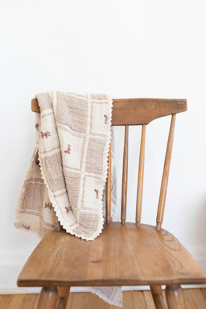 A brown wooden chair with the Organic Cotton Baby Blanket - Pony, featuring a beige and brown pattern, draped over its backrest, set against a plain white wall and wooden floor.