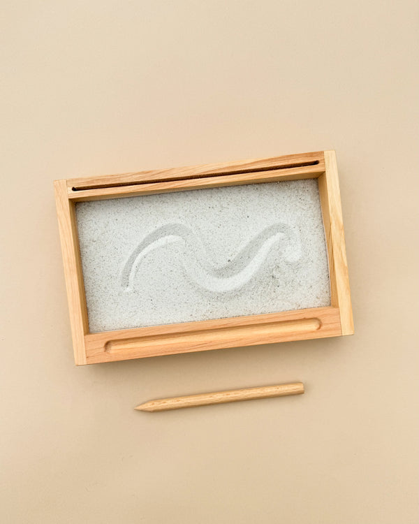 A sand tray with a flash-card holder features a wavy line drawn in the sand. Below, a wooden stylus used for this tactile learning experience rests on a beige background. The tray has a wooden frame with a slot designed for storing the stylus, enhancing writing skills through play.