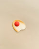 A minimalist image featuring a wooden, kidney-shaped dish on a beige background. A single round object, the Erzi Wooden Sunny-Side Up Egg, rests on the dish, creating a simple yet striking contrast.