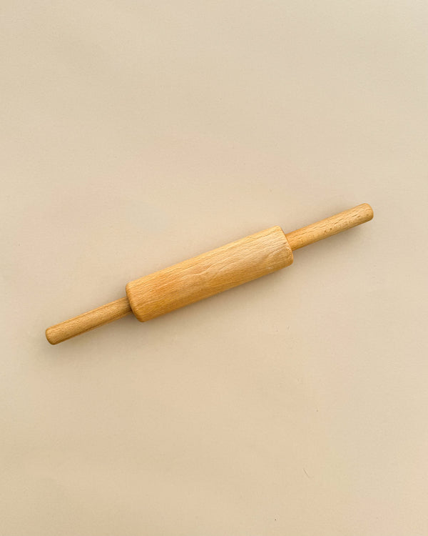 A Handmade Wooden Pretend Rolling Pin with handles on both ends, photographed against a plain, light beige background. The heirloom-quality rolling pin, crafted from maple beech wood, features a smooth finish and a typical cylindrical shape used for rolling out dough.
