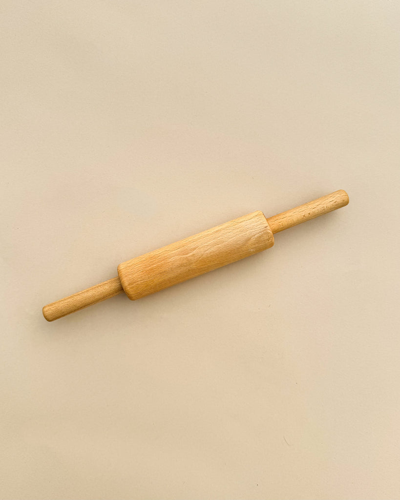 A Handmade Wooden Pretend Rolling Pin with handles on both ends, photographed against a plain, light beige background. The heirloom-quality rolling pin, crafted from maple beech wood, features a smooth finish and a typical cylindrical shape used for rolling out dough.