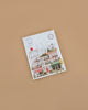 The Paint By Number Kit - New York features a charming illustrated cover with whimsical pastel buildings and shops on a light brown background, evoking the creative spirit of cityscapes and the fun of paint-by-number sets.