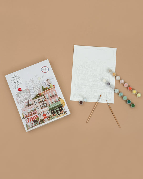 The Paint By Number Kit - New York includes a cityscape box, outlined painting sheet, two brushes, and paint pots—everything you need to unleash your creativity and create a masterpiece on a beige surface.
