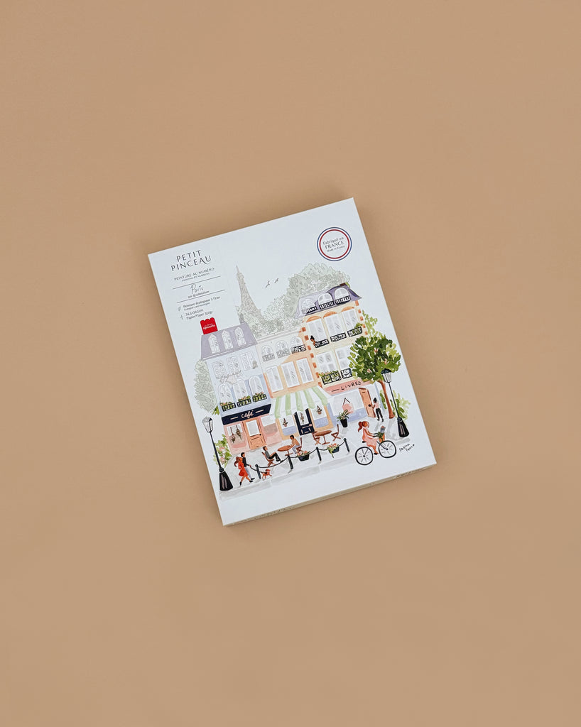 The Paint By Number Kit - Paris features an illustrated cover of a vibrant Paris street scene filled with color and creativity, placed on a plain tan background—ideal inspiration for aspiring artists.