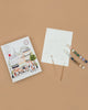 The Paint By Number Kit - Paris includes a partially outlined coloring sheet of a Parisian street scene, two paintbrushes, and a colorful palette—ideal for aspiring artists ready to create on a beige surface.