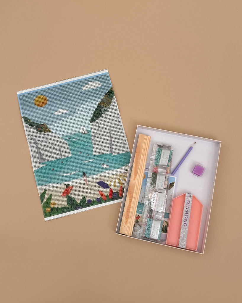 The Diamond Painting Kit - Stiniva Beach by Maja Tomljanovic displays a coastal scene with cliffs, sea, sailboats, and beachgoers. The kit includes diamond beads, tools, labeled packages, and an adhesive cotton canvas.