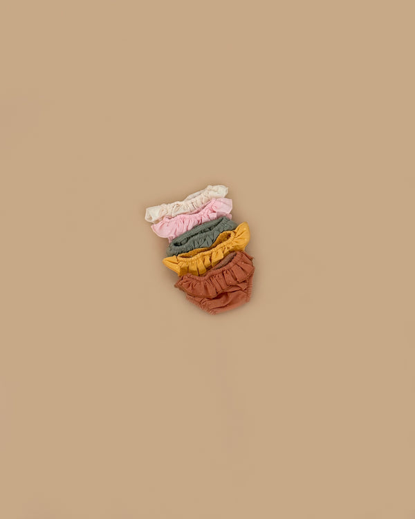 The Minikane Doll Clothing | Cotton Ruffled Underwear Set includes five pairs in cream, pink, green, mustard yellow, and brown, shown stacked on a beige background.