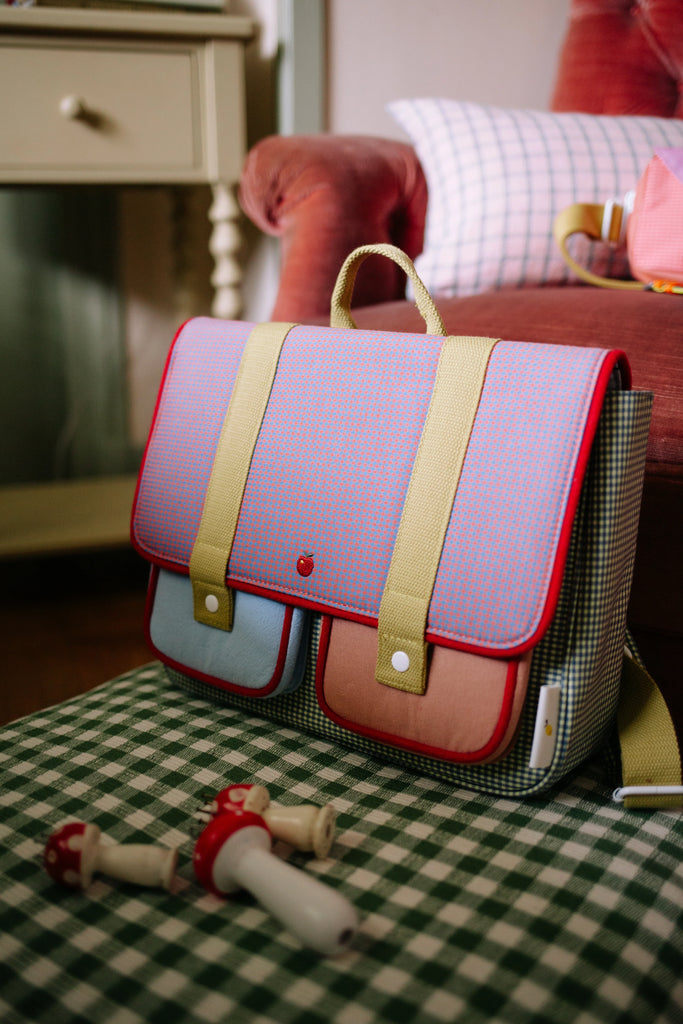 The Sticky Lemon School Bag | Lemon Tree | Gingham Midnight + Moon, featuring yellow straps and adjustable handles, rests on a green checkered surface beside wooden toys, in front of a red armchair and side table.