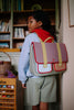 A child wearing a blue hoodie and plaid shorts stands indoors with the Sticky Lemon School Bag | Lemon Tree | Gingham Midnight + Moon, featuring adjustable shoulder straps, looking over their shoulder in a room with bookshelves.