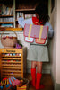 A child with a Sticky Lemon School Bag | Lemon Tree | Gingham Midnight + Moon and red knee-high socks stands by a bookshelf, reaching for a book, with baskets of yarn and a wooden cabinet of sewing threads nearby.