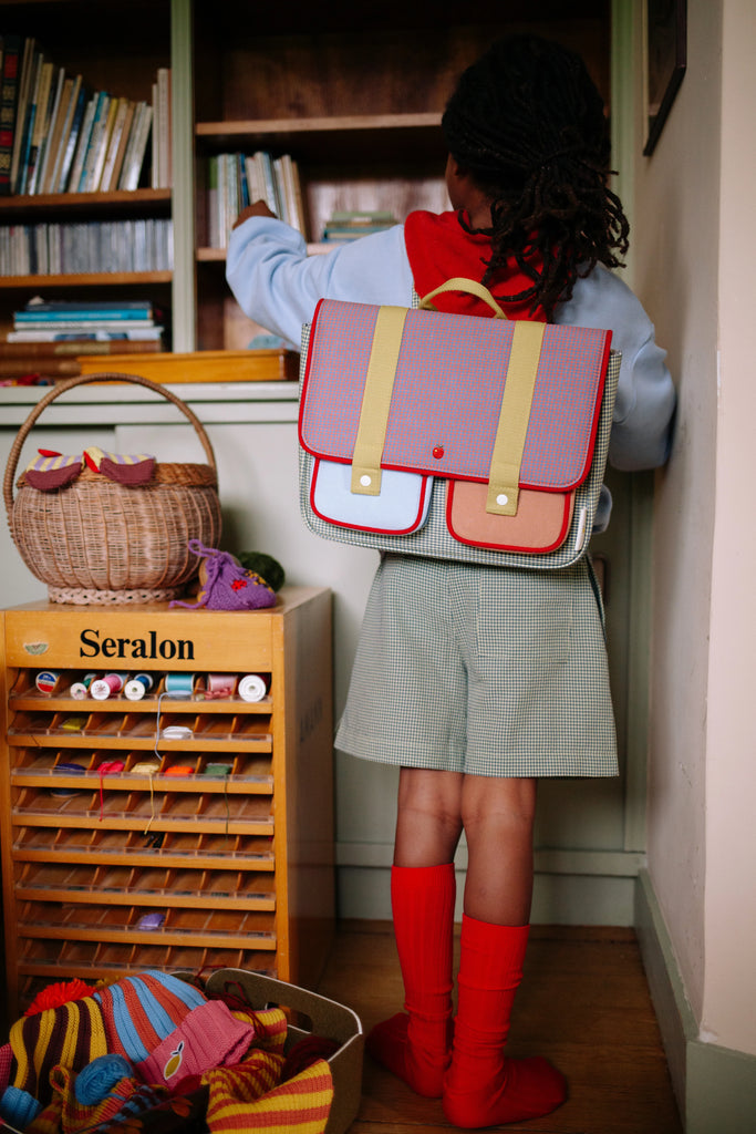 A child with a Sticky Lemon School Bag | Lemon Tree | Gingham Midnight + Moon and red knee-high socks stands by a bookshelf, reaching for a book, with baskets of yarn and a wooden cabinet of sewing threads nearby.