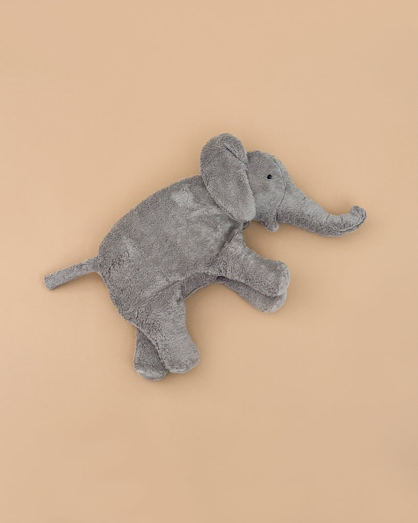 The Senger Naturwelt Cuddly Animal - Elephant is a soft, gray plush toy made from organic cotton with floppy ears, a long trunk, and small black eyes, lying on a plain light brown background. It doubles as a comforting warming pillow.
