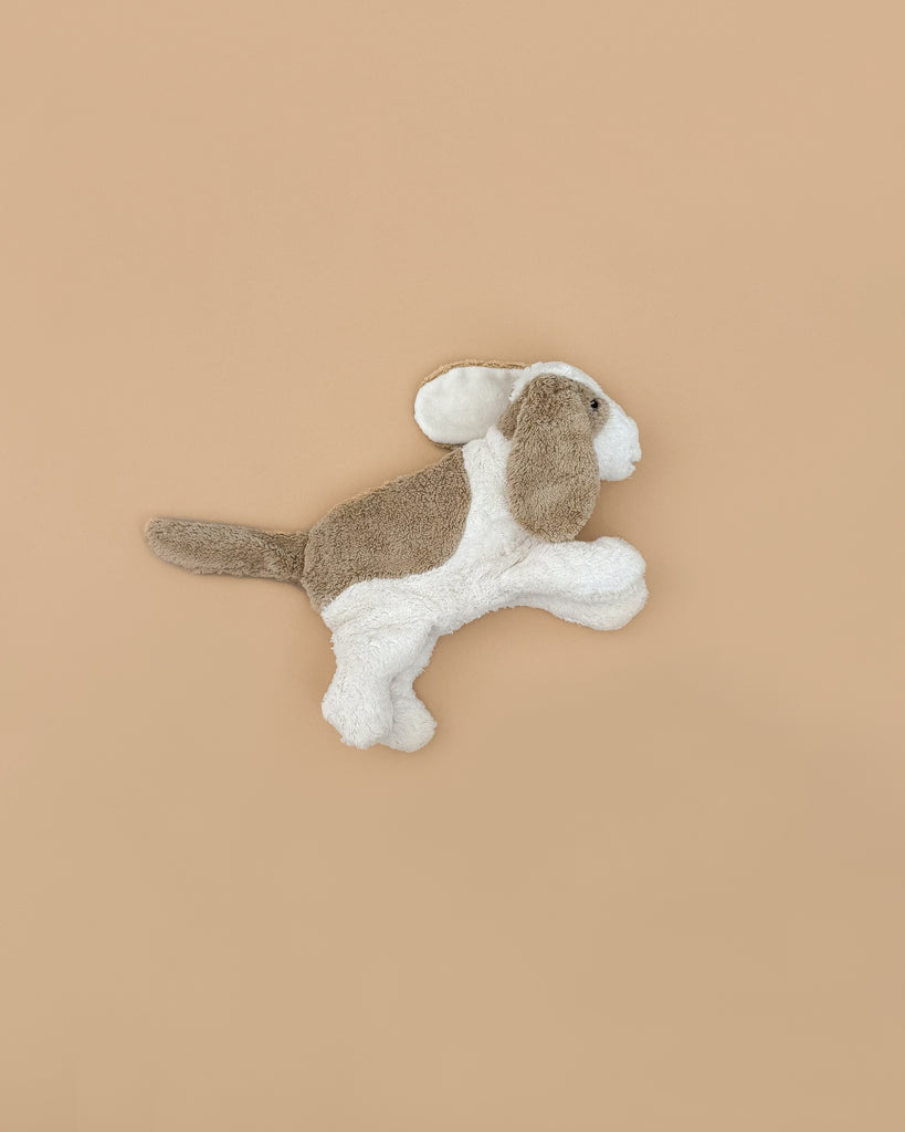 The Senger Naturwelt Cuddly Animal - Dog features soft brown and white fur, floppy ears, and a long tail, shown lying on a plain light brown background.