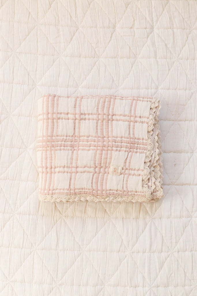 The Organic Cotton Baby Blanket - French Plaid, Petal, featuring pale pink and cream plaid and lace trim, is folded on a white diamond-quilted surface.