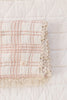 The Organic Cotton Baby Blanket - French Plaid, Petal features a folded, cream-colored fabric with a pink plaid design and delicate lace trim, displayed on a quilted white surface.