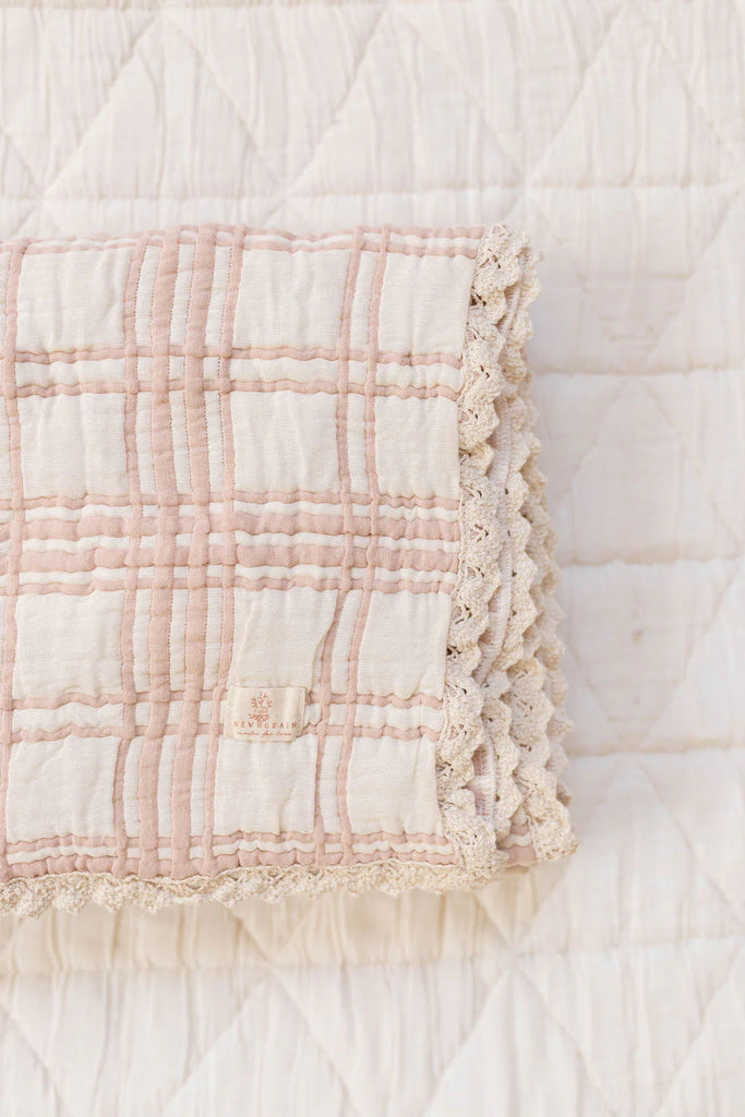 The Organic Cotton Baby Blanket - French Plaid, Petal features a folded, cream-colored fabric with a pink plaid design and delicate lace trim, displayed on a quilted white surface.