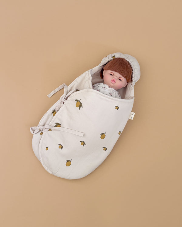 A lifelike baby doll with straight brown hair, dressed in white, lies on a plain light brown background wrapped in the Konges Sloejd Doll Sleeping Bag - Lemon, made from organic cotton with a lemon print.