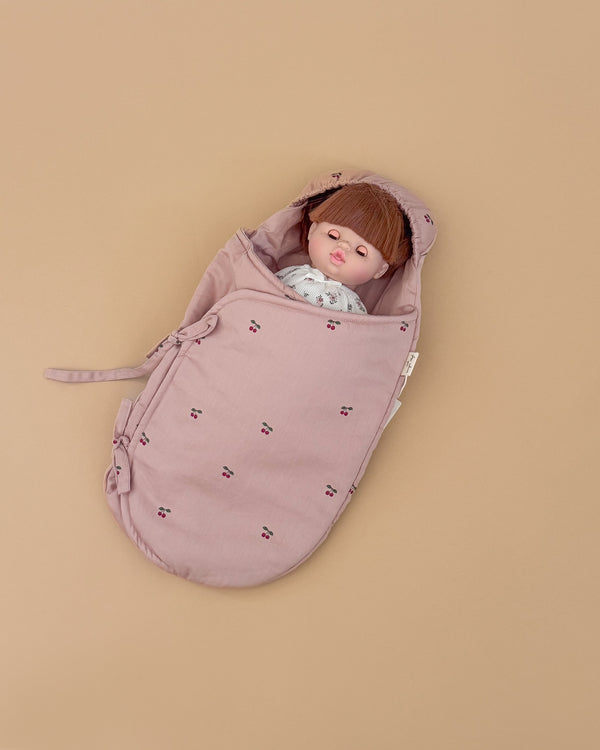 A doll with straight brown hair and bangs lies on a plain beige background, wrapped in the Konges Sloejd Doll Sleeping Bag - Cherry Blush, made from organic cotton with pink patterns and small embroidered details.