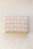 The Organic Cotton Twin Size Quilt - Meadow, Petal features a pastel pink and white checkered pattern with blue floral accents, made in Portugal, and is shown folded on a cream diamond-quilted surface.