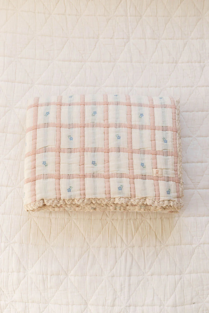 The Organic Cotton Twin Size Quilt - Meadow, Petal features a pastel pink and white checkered pattern with blue floral accents, made in Portugal, and is shown folded on a cream diamond-quilted surface.