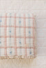 The Organic Cotton Twin Size Quilt - Meadow, Petal features a pink and white checkered pattern with small blue floral accents and lace trim, made in Portugal, folded neatly on a white textured surface.