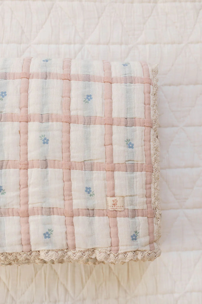 The Organic Cotton Twin Size Quilt - Meadow, Petal features a pink and white checkered pattern with small blue floral accents and lace trim, made in Portugal, folded neatly on a white textured surface.