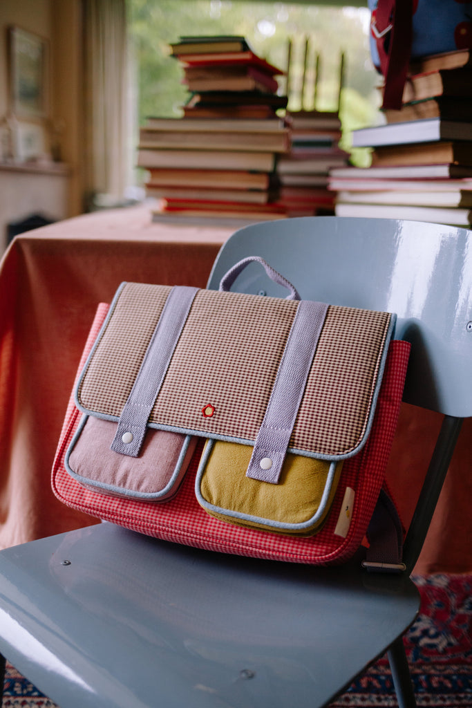 A Sticky Lemon School Bag | Lemon Tree | Gingham Flower + Rose, crafted from waterproof nylon and recycled PET bottles with pink, yellow, and beige pockets, sits on a gray chair; stacked books are visible on a table in the background.
