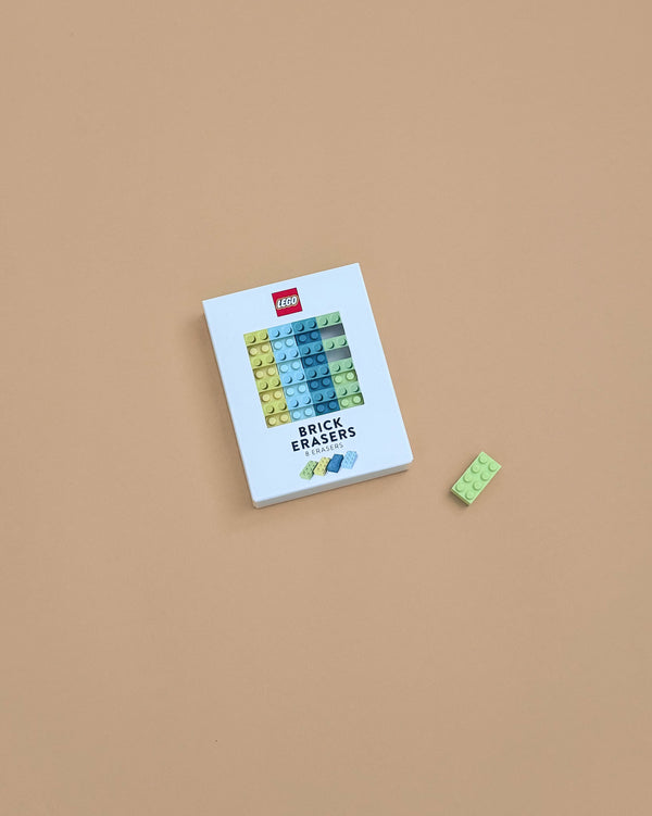 A white box labeled "Lego Brick Erasers" sits on a beige surface, featuring colorful erasers shaped like LEGO bricks. A green brick-shaped eraser lies beside the box, making this set a fun party favor for LEGO fans.