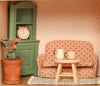 The Maileg Fully Furnished SuitCasa Set features a miniature living room with a polka-dot sofa, small wooden table with two mugs, potted plant, green cabinet with alarm clock, and patterned wallpaper in striped and floral designs.