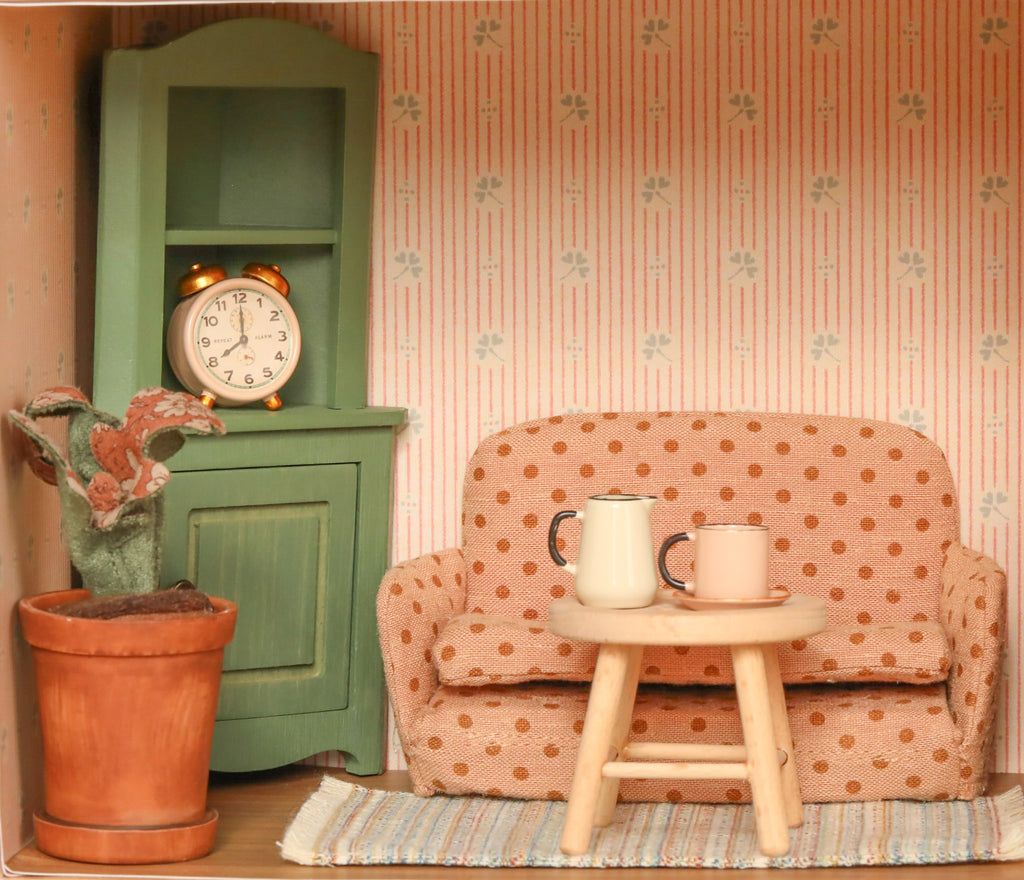The Maileg Fully Furnished SuitCasa Set features a miniature living room with a polka-dot sofa, small wooden table with two mugs, potted plant, green cabinet with alarm clock, and patterned wallpaper in striped and floral designs.