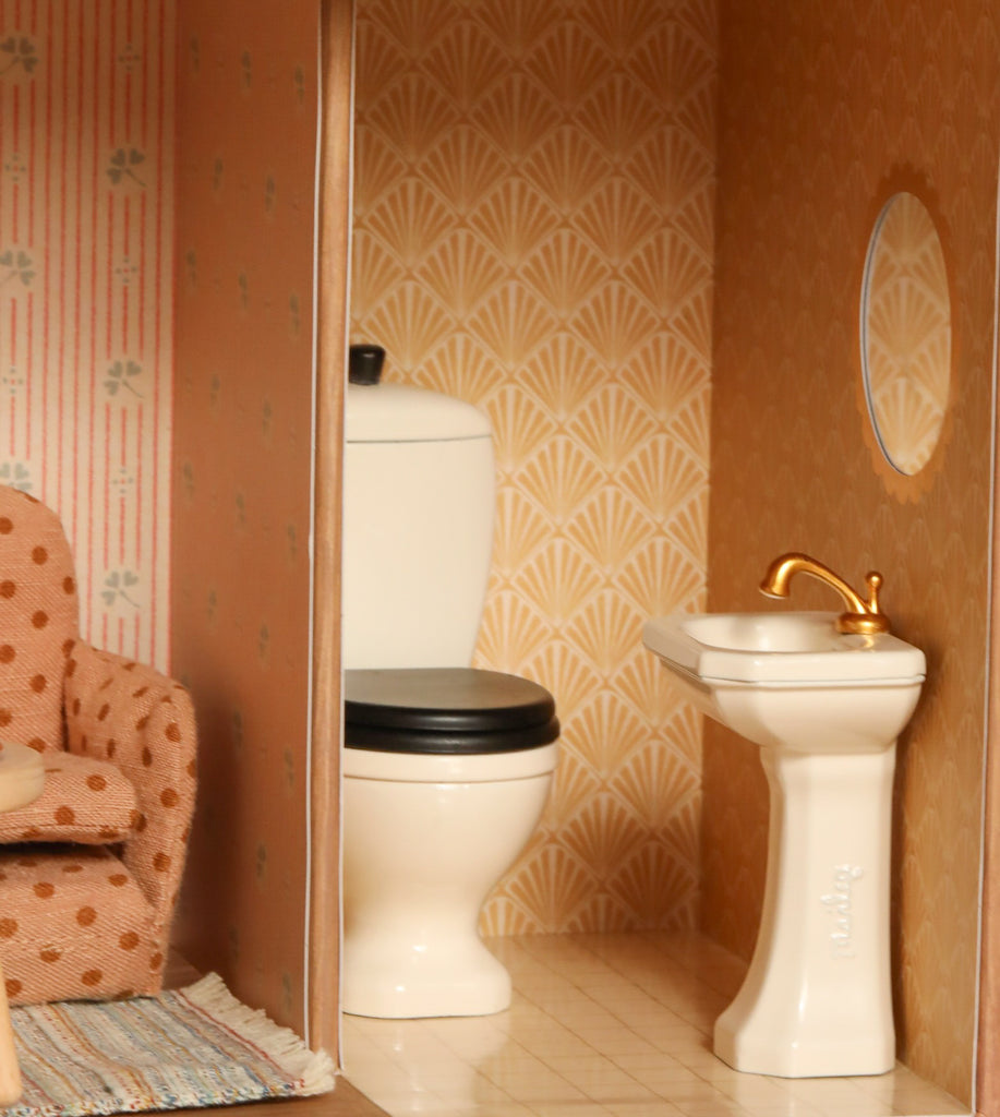 The Maileg Fully Furnished SuitCasa Set includes a miniature bathroom with a white toilet with black seat, pedestal sink with gold faucet, round mirror, patterned wallpaper, plus a polka-dotted chair and rug.