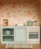 The Maileg Fully Furnished SuitCasa Set features a miniature vintage kitchen with a green mug, pot, can of tomatoes, small oven, woven rug, and kitchen counter—ideal for any Maileg mouse house with its pink floral wallpaper backdrop.